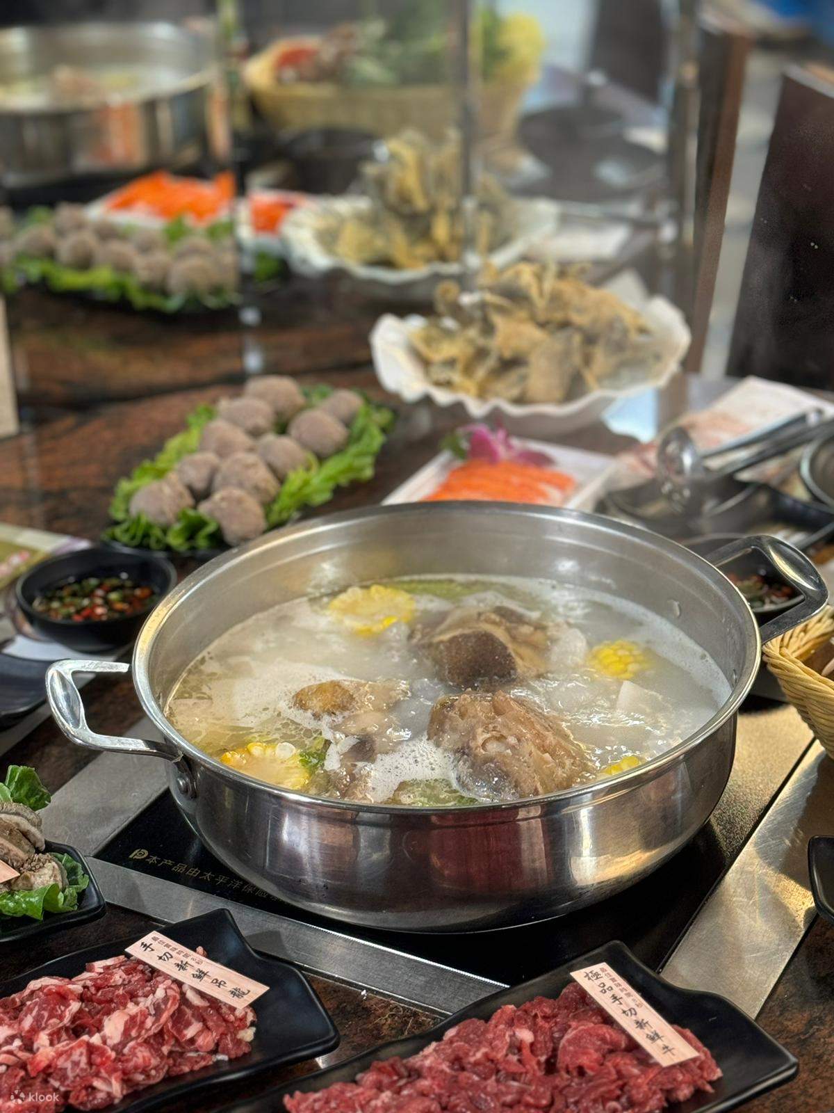 A steaming hot pot filled with meat, corn, and vegetables sits at the center of a table surrounded by plates of raw meats, vegetables, and seafood, reflecting the vibrant and authentic dining culture of Hong Kong's Treasure City Hot Pot in Mong Kok.